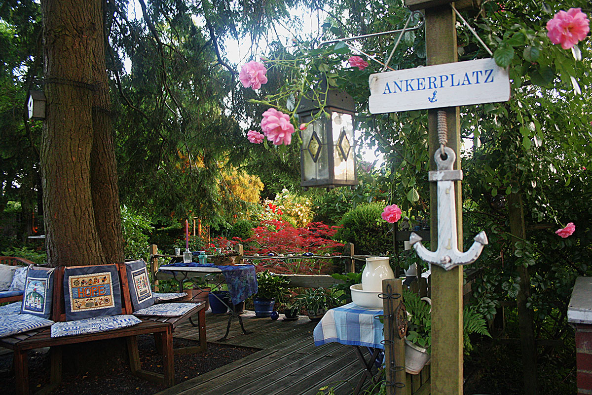 Maritime Sitzecke mit Schild „Ankerplatz“, Laterne und rosa Kletterrosen unter einem großen Baum – Foto von Karin, Platz 2 beim Gärtner Harry Fotowettbewerb „Dein schönster Garten-Moment im Sommer“